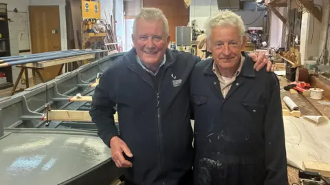 Two men in overalls stand next to the vessel they have been painting and sanding