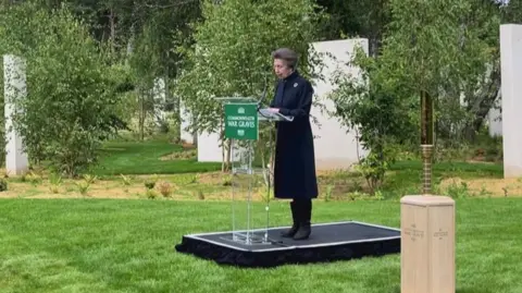 A woman in a dark  coat and boots stands on a podium surrounded by trees and large upright stone slabs.