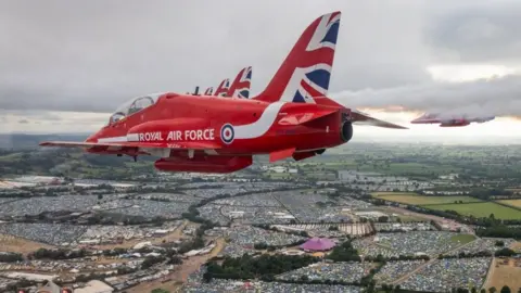 A Red Arrow is flying over the Glastonbury Festival, which is sprawling over multiple fields.