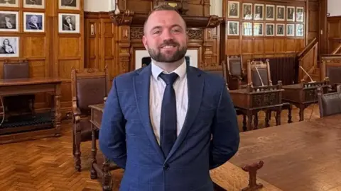 EMMA DRAPER Devon Watson, a man with a beard and short brown hair, he wears a navy sui, and has a bright smile as he stands in the Douglas Council; chamber.