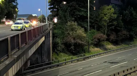 Police are parked along a bridge, with a three lane motorway underneath. It is empty except for a damp splatter on the road