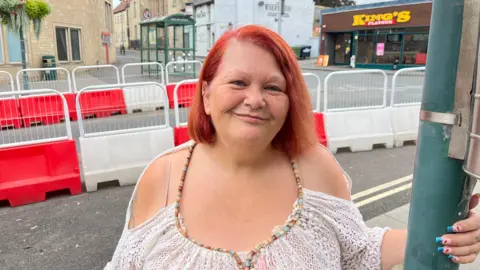 Chairman of Marden Community Hub Dawn Perkins with red hair and a white beaded top smiling and standing in front of red and white plastic barriers on the high street in Calne.