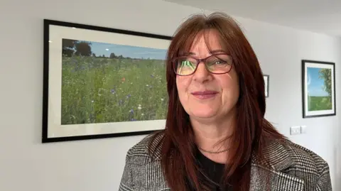 A woman with long brown hair smiles as she stands in a white-walled room in front of a painting of a meadow full of wild flowers. She is wearing scarlet-coloured glasses and a black and white jacket with a fine check pattern.
