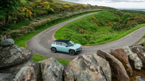 TimbuktuContent.com/EaseeSunRun A small blue car drives down a single track country road.
