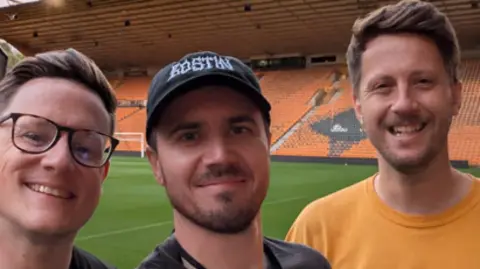 The trio are inside Wolves' ground when it is empty and are stood together for a selfie. Kieran has glasses on a brown hair, Nathan has a black on a short beard and Alex has short styled brown hair and is wearing an orange T'shirt. All three are smiling at the camera.