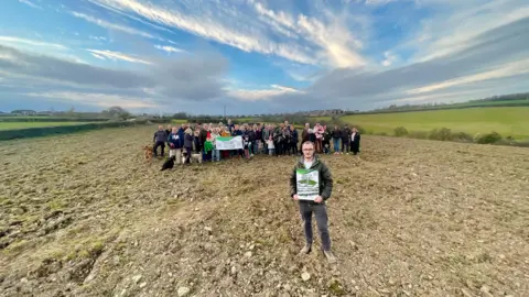 A man in sunglasses, a green puffer jacket and grey t-shirt is standing in a field which dips into a valley. Behind him are forty-two adults, six children and seven dogs. Four members of the group hold a banner which reads ‘Save S12 Green Belt’. In the distance is another field coming up out of the valley. There are trees, a school and a bridleway on the edge of the fields. The sun is setting.