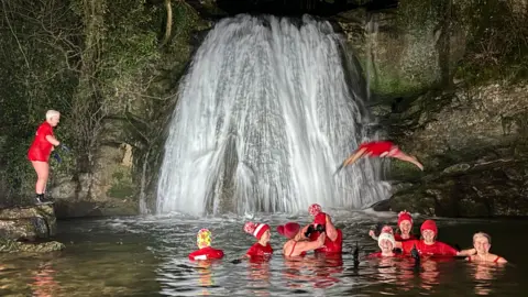 There is a large waterfall in the background with people in the pool in front of it wearing red hats and t-shirts