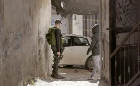 A Palestinian fighter with a rifle by his side, standing in an alleyway