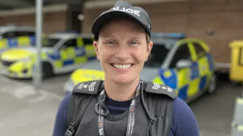 Detective Sergeant Jen Jeffcoat-Marsh smiling wearing a police baseball cap, stab vest and sergeant rank slides