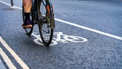 Getty Images A cyclist riding in a designated bike lane marked by white lines and a painted bicycle symbol on a road. Only the bike’s rear wheel and the rider’s legs and shoes are visible.