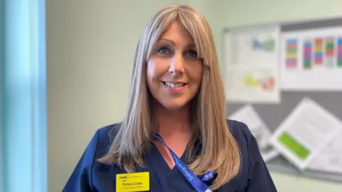 Teresa Cope has long blonde straight hair with a parted fringe. She is smiling and wearing dark blue scrubs with a blue lanyard around her neck and a yellow name badge that says "hello my name is Teresa Cope, chief executive officer". She is standing in an office with a pinboard on the right.