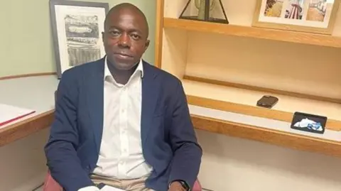 Bayo Abala Man wears a white shirt and navy jacket sits on an armchair with a clipboard on his lap. He has framed photographs on shelving behind him.