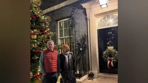 Stephen Reynolds A man and a woman are standing in front of the door to 10 Downing Street. There is a large fir Christmas tree to their left, covered in decorations. There is a large wreath on the door of the building, with a red and white ribbon trailing down from it