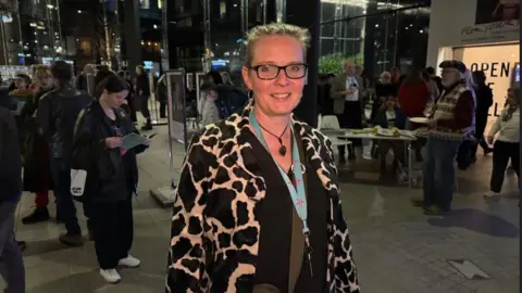 Phil Chapman / BBC A woman with blonde hair, tied back, wearing glasses and a cow print coat, smiles at the camera as she stands in an auditorium 