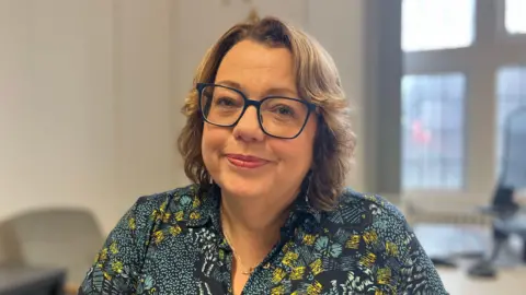 Wendy Thomas, Head of Neurodivergence Service Wales. She has brown hair with highlights, and is wearing dark blue spectacles and a navy and mustard patterned shirt. 