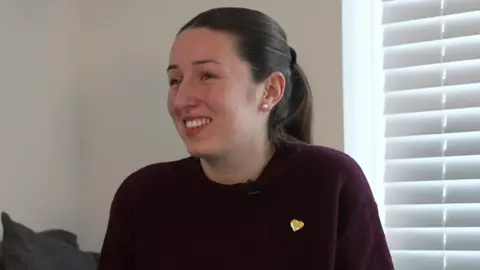 Young woman, 25, wearing a burgundy top with chestnut brown har tied back. She is wearing a yellow heart pin badge on her top.