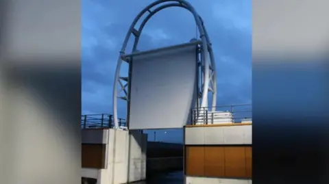 Rotherham Council A large barrier which consists of a rectangular gate suspended vertically between two steel arms that form an arch-like frame. The gate appears to be raised, revealing water below. On either side of the barrier are concrete walls topped with railings.