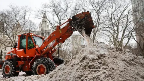 An orange plow dumps snow onto a large, grey snow pile in New York City. City Hall can be seen in the background.
