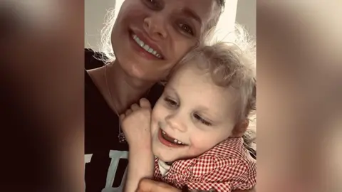 Family photograph Sarah (left) and her young daughter Isla hugging. Sarah has blond hair tied back and is wearing a burgandy and white t-shirt with Isla who has blond curly hair and weating a red and white gingham top. They are both smiling. 