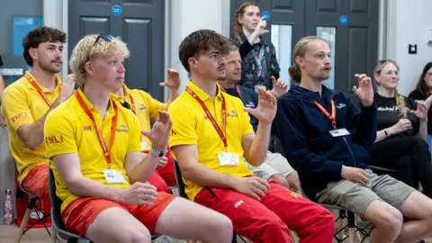 Six men and one woman are sitting while a woman stands, all looking forward. They are all curling their left hands in the C letter BSL sign. Half of them are wearing RNLI lifeguard uniforms and the others The Deaf Academy uniforms. All of them are wearing halyards. Behind them is a set of doors.