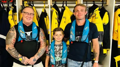 Dai Jones, Marley and Marley's dad stand together in Barry Dock RNLI station in front of the crew member's yellow waterproof jackets. Dai wears glasses and a black vest, and has tattoos on both arms, Marley has short brown hair and wears a grey pattern football shirt, and Joe has short light brown hair and wears a black t-shirt and jeans. All wear blue RNLI life vests. 
