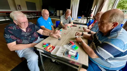 MARTIN GILES/BBC A group of six men sit round a table at the working man's club playing cards. They have counters that they are using to place bets. They are largely retired and are wearing t-shirts or light summer shirts