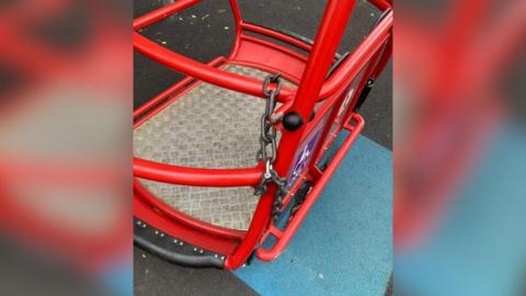 A red children's play swing, with a disability logo on it, is padlocked with a chain.