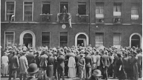 A black and white photo of a crowd facing a building in London. A man stands at a balcony on the first floor, looking back at the crowd. 