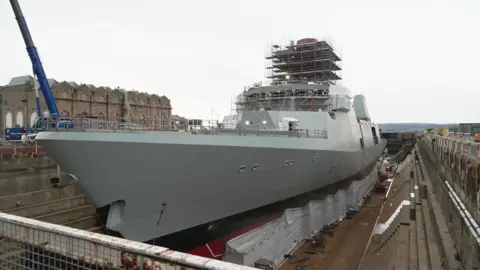 HMS Venturer a large grey warship with black and red paint on the underside. It is in a dry dock with cranes and other machinery around it. The sea is visible in the distance.
