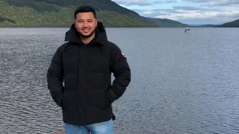 Man in a black jacket and jeans with black hair and beard stands smiling on a jetty in front of a lake with hills and a boat beyond.