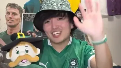 A man in a green outfit and bucket hat waves and holds a Plymouth Argyle mascot.