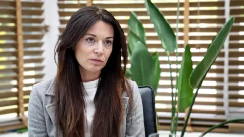 Zoe Trafford has long brown hair and wears a grey blazer and white top underneath. She is sitting against a background of wooden blinds against windows. The leaves of an indoor plant are also there.