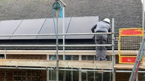 A man dressed in grey with a white hard hat is turned away from the camera, standing on scaffolding and working on a solar panel, which is fixed to the roof of a bungalow. 