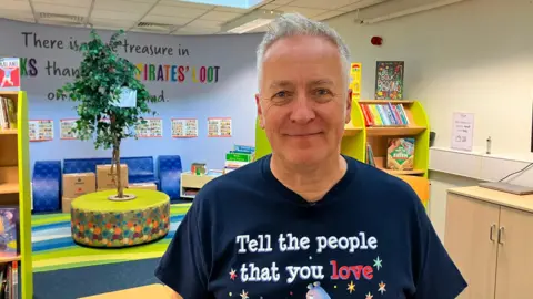 A man with silver hair stands in a library at a primary school and smiles at the camera. He wears a navy blue shirt that says "tell the people that you love" at the top.