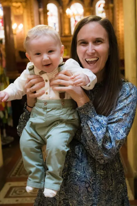Kimber Kreative Photography A mum standing in the aisle of a Victorian chapel holds her baby boy up to the camera, both are smiling. 