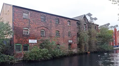 Ian Hughes A large brick building alongside a canal with plants growing up the walls and a number of arched windows set in the wall