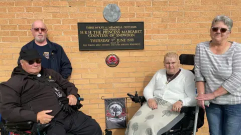 Four elderly people, two using wheelchairs stand and sit infront of a plaque on a wall