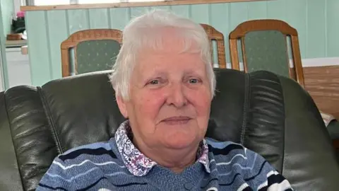 An older woman with grey hair smiles at the camera, sitting on an armchair. She is wearing a blue and white jumper, with a patterned pink collar.