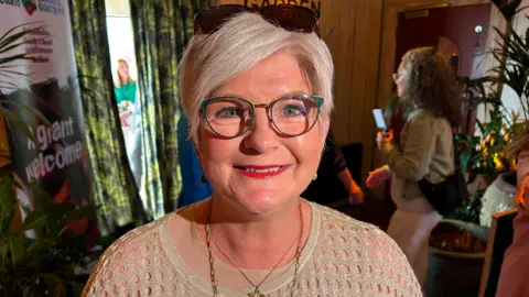 A woman in a light brown jumper wearing necklaces. She has white short hair and wears glasses and red lipstick. There are sunglasses on her head.