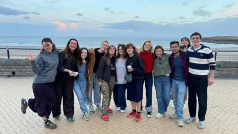 IJPR Ten woman and two men are smiling while standing by the sea on a pavement at Barry Island. Ruth Jones is in the centre, with Ella Bruccoleri to her left. 