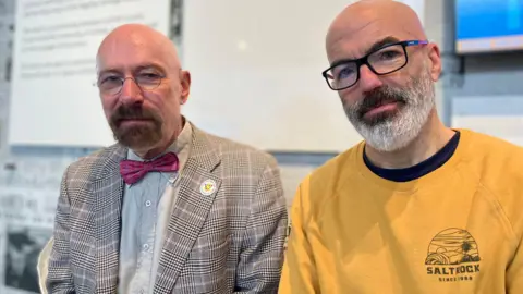 Jon Wright/BBC Rainer Schulze and Mark Curtis sit next to each other looking solemnly at the camera.