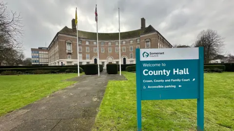 County hall in Taunton, a large four-storey brown brick building with three flags on tall flagpoles flying in front of it, and grey cloudy skies behind. A sign in the foreground reads 'Welcome to County Hall'.