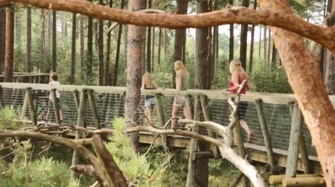 Four people including at least two children walk along an elevated pathway, enclosed by wire mesh up to hip height and surrounded by trees.