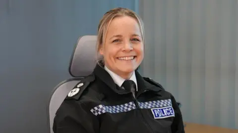 Katy Barrow-Grint is sitting in a desk chair in police uniform. She has long blonde hair and is smiling.