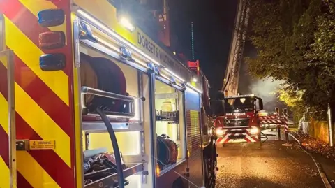 Dorset & Wiltshire Fire & Rescue Service fire engines along a street in the dark, lights on, equipment being used form the sides and top