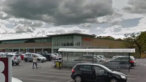 Google A car park at the Sainsbury's store on Wilmslow Road, Cheadle, with cars parked up and shoppers pushing trolleys.