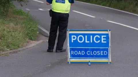 A blue sign of a road closure saying 'police, road closed', with an officer is black trousers stood close by.