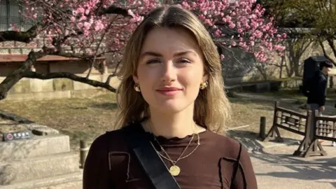 Saoirse Cleary Saoirse Cleary, smiling, standing in front of a tree with pink blossom.