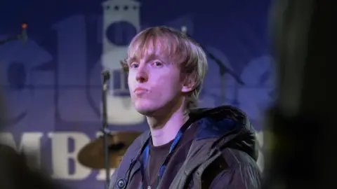 Ioan Bowen-Pickett who has blonde hair with a long fringe, wearing a black hooded jacket and stood on a stage with a drumkit behind him. He is looking off to the side with a serious expression on his face.