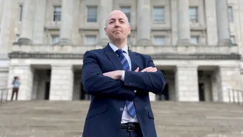David Young/PA Wire Jon Burrows. a man with a shaved head, stands on the steps of Stormont with his arms folded, looking down at the camera. He is wearing a navy suit, a white shirt and a navy and mid-blue striped tie. 